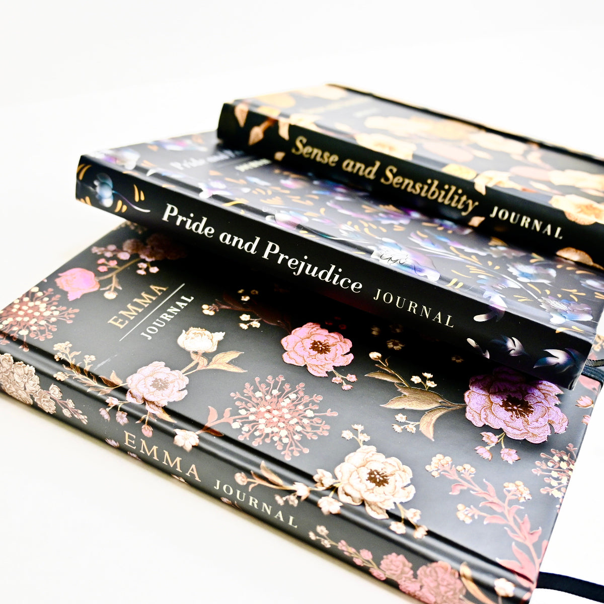 Three black journals with floral designs are in fan shape. The journals have the titles "Emma Journal", Pride and Prejudice Journal" and "Sense and Sensibility Journal".