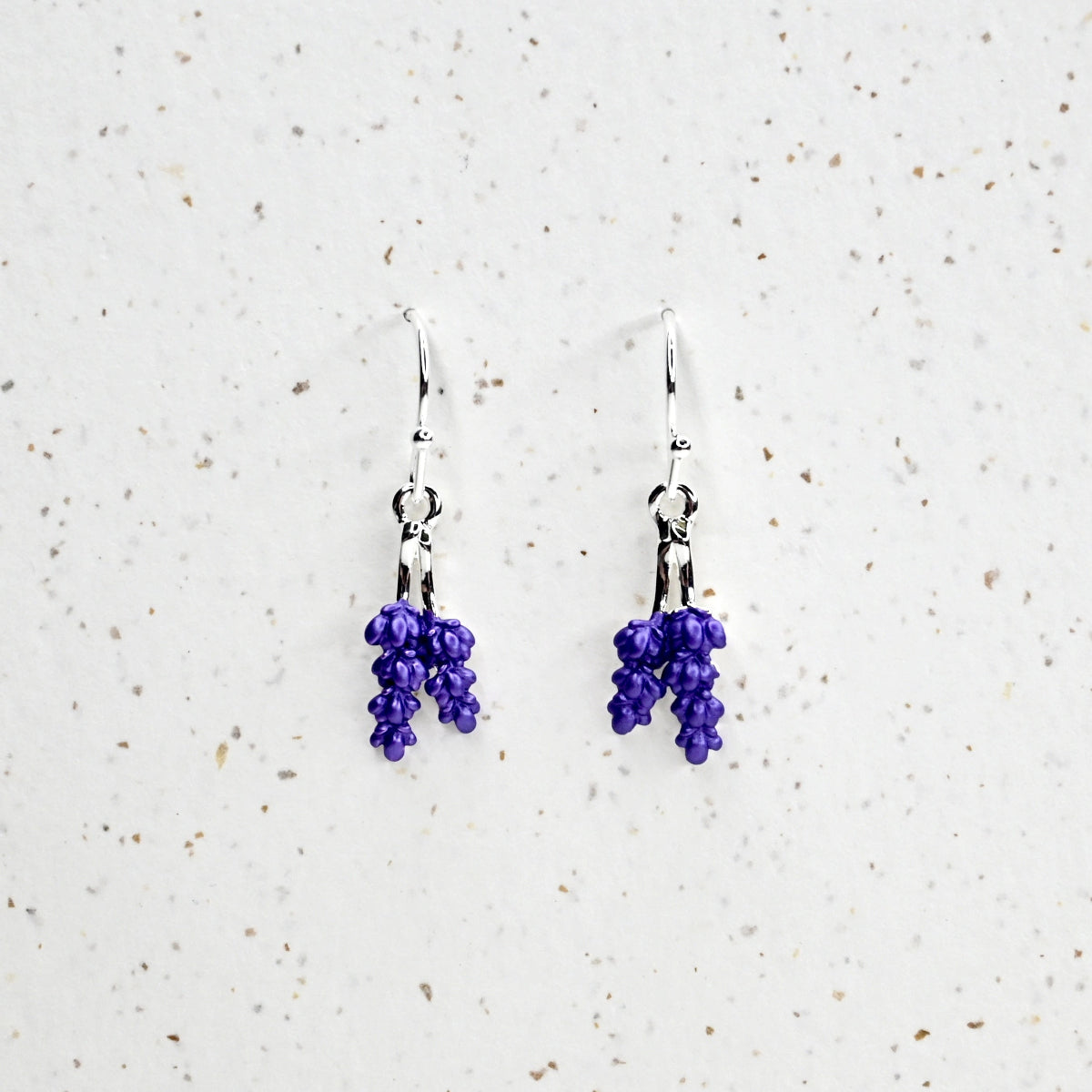 A pair of vibrant purple lavender earrings displayed against a white and silver earring stand, set against a textured white background.