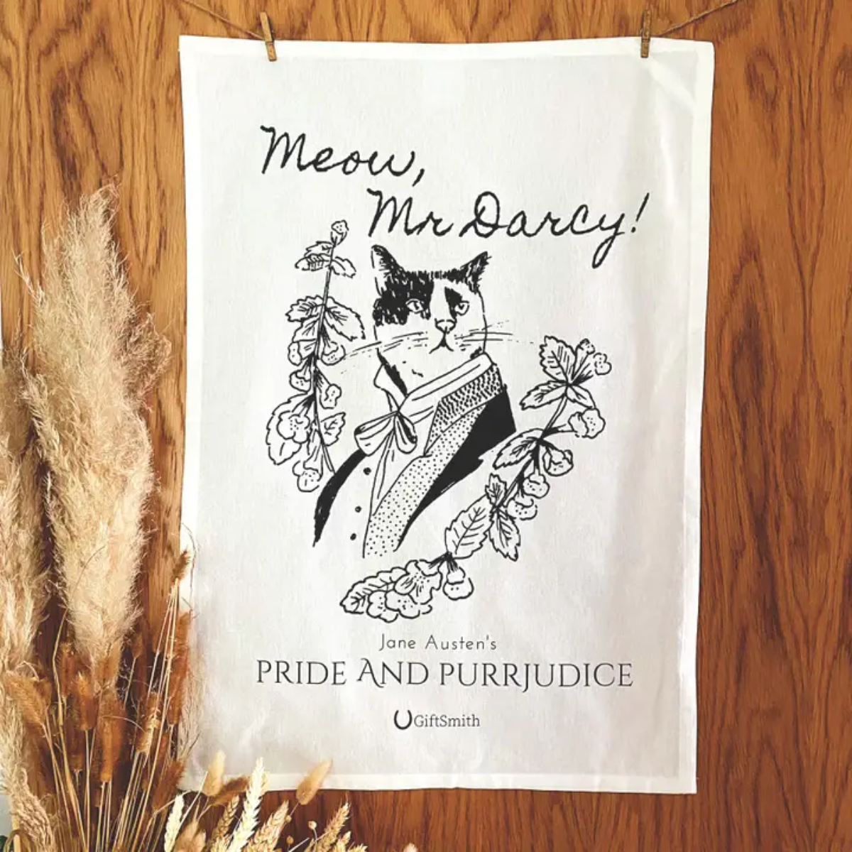 Tea towel showing a line drawing of a cat dressed as Mr Darcy, the towel is hanging from a clothes line in front of a wood effect background with bunch of dried grasses in the foreground.