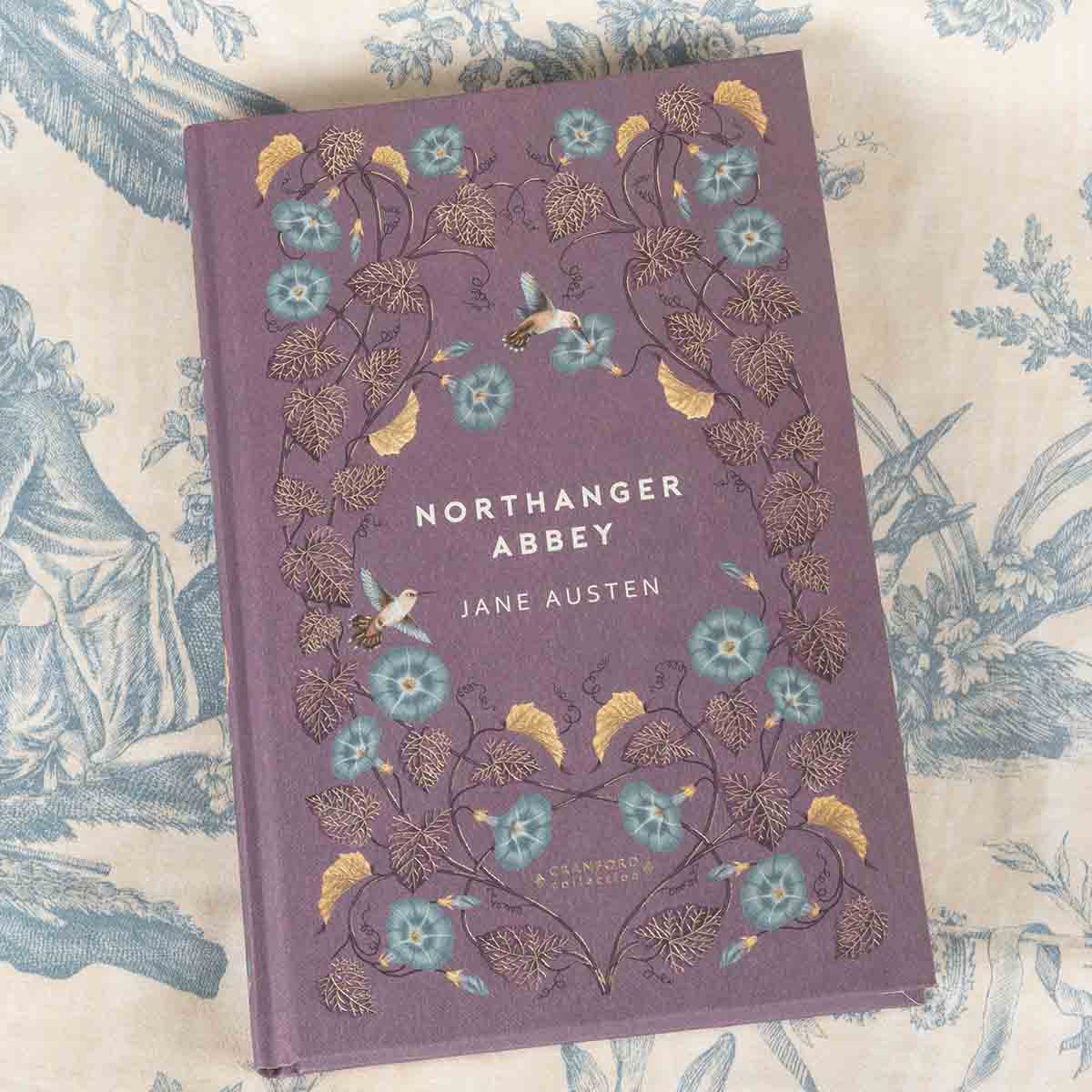 Northanger Abbey front cover - decorated in blue lilies, gold leaves and branches, and hummingbirds. The background colour is a purplish-pink.
