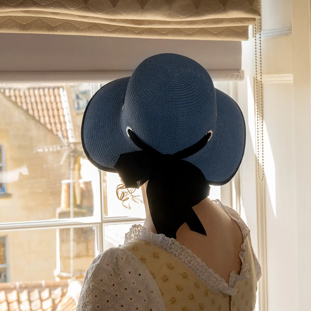 photograph of model looking out of a window while wearing blue open-back regency bonnet