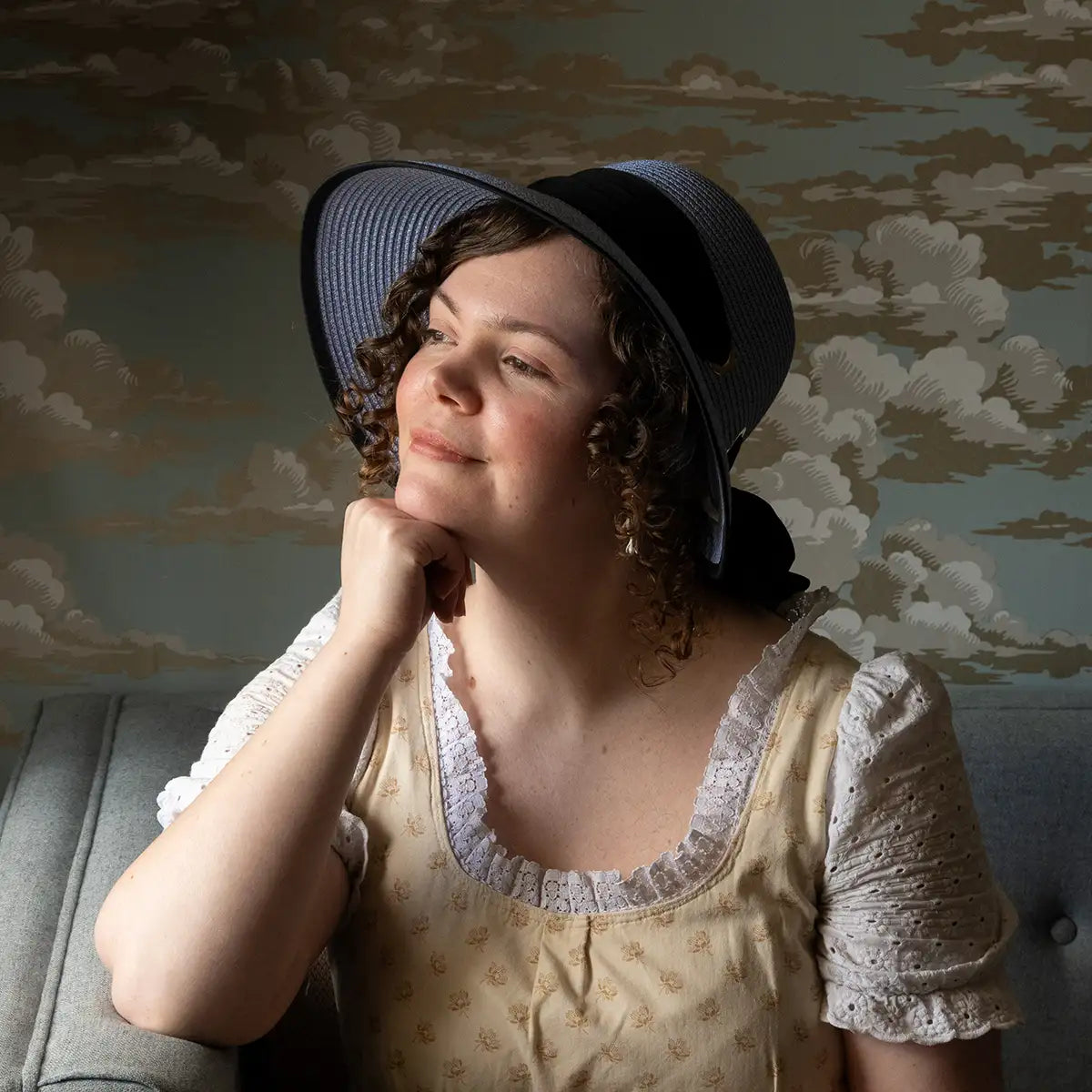 A model in a regency dress wearing a blue open-back regency bonnet against a blue background with gold clouds