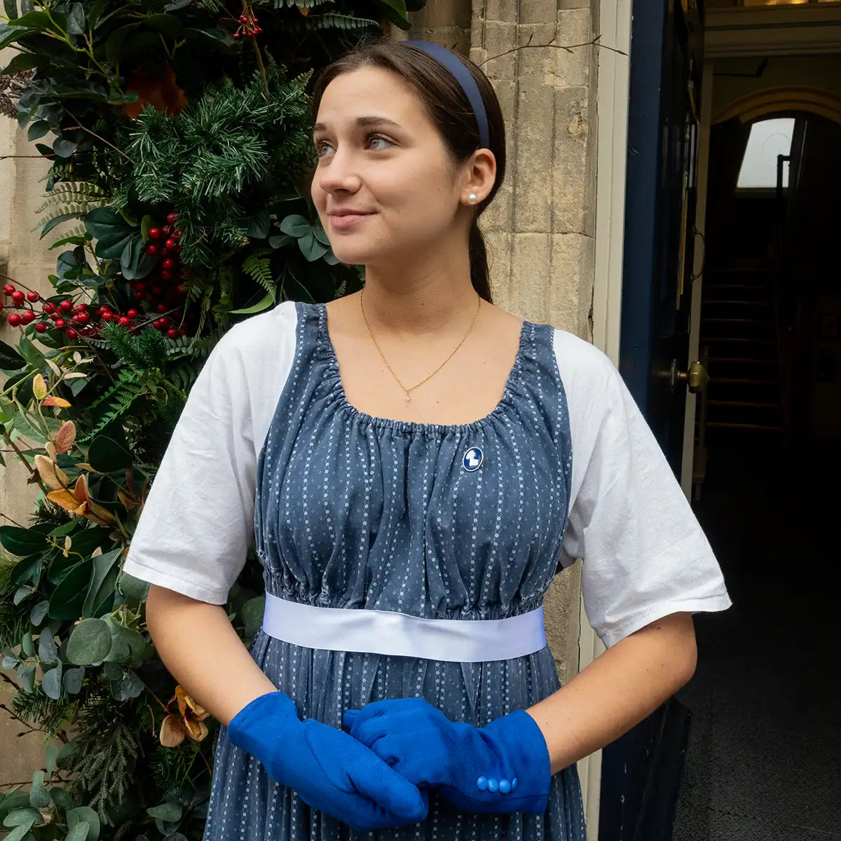 Woman in a blue dress with white sleeves and blue gloves standing in front of a stone building with greenery.