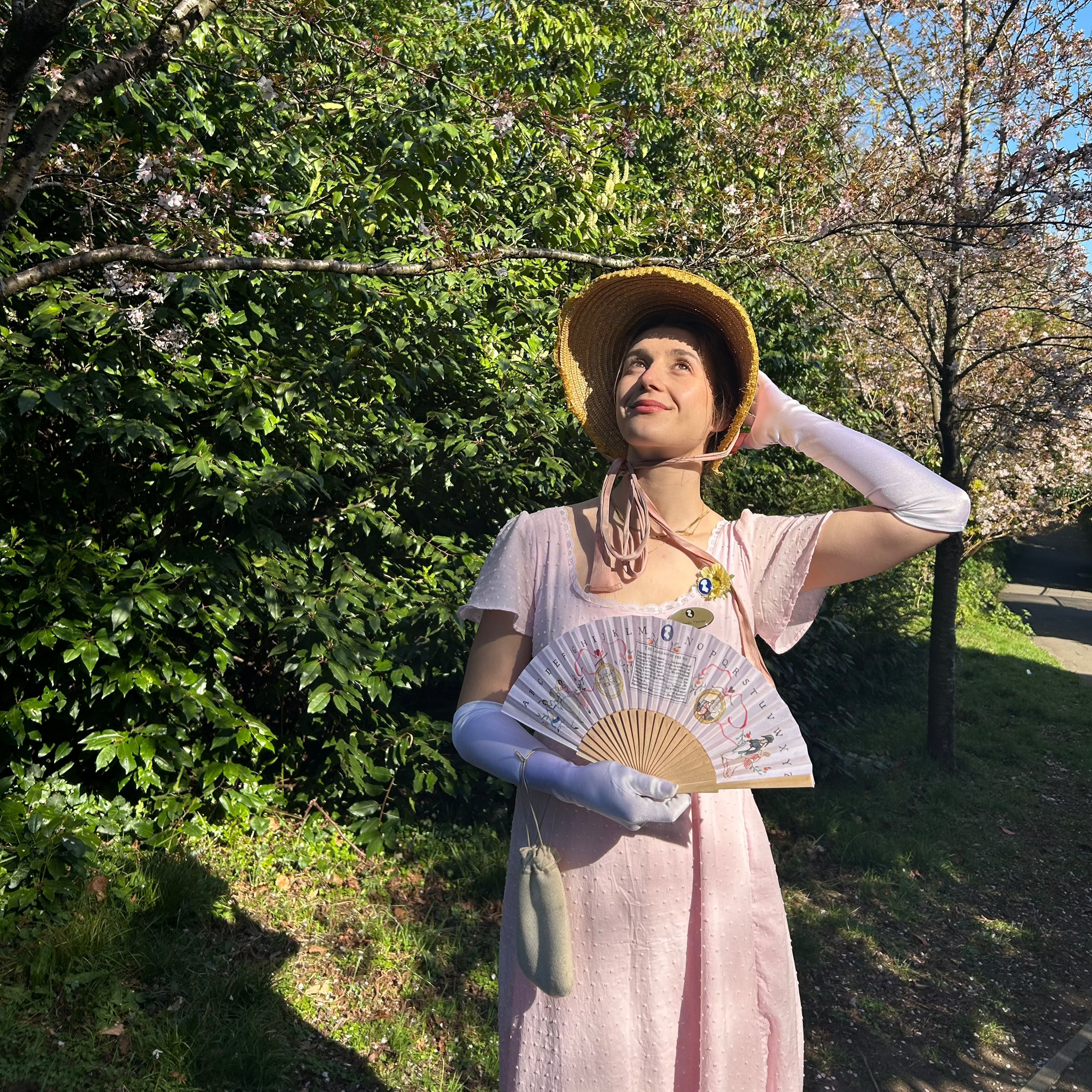 Isabella Thorpe from The Jane Austen Centre stands on a grassy pathway surrounded by cherry blossom trees, with a pale pink, patterned, Regency fan in her hand.