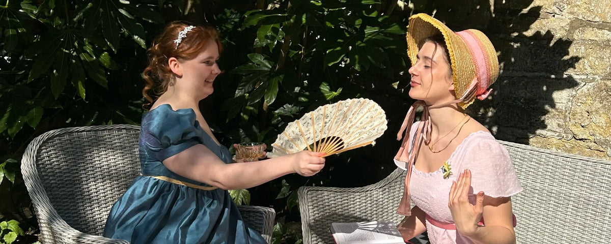 2 Women in regency dress and a pinci. One woman is fanning the other