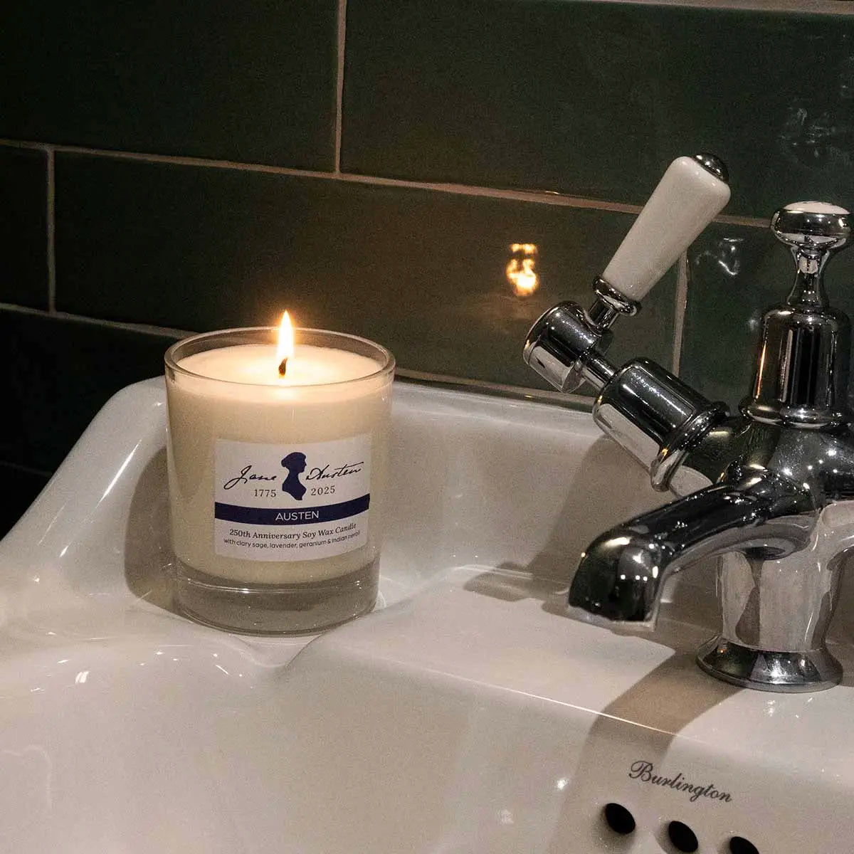 candle in bathroom on a sink against a green tile wall next to silver and white taps