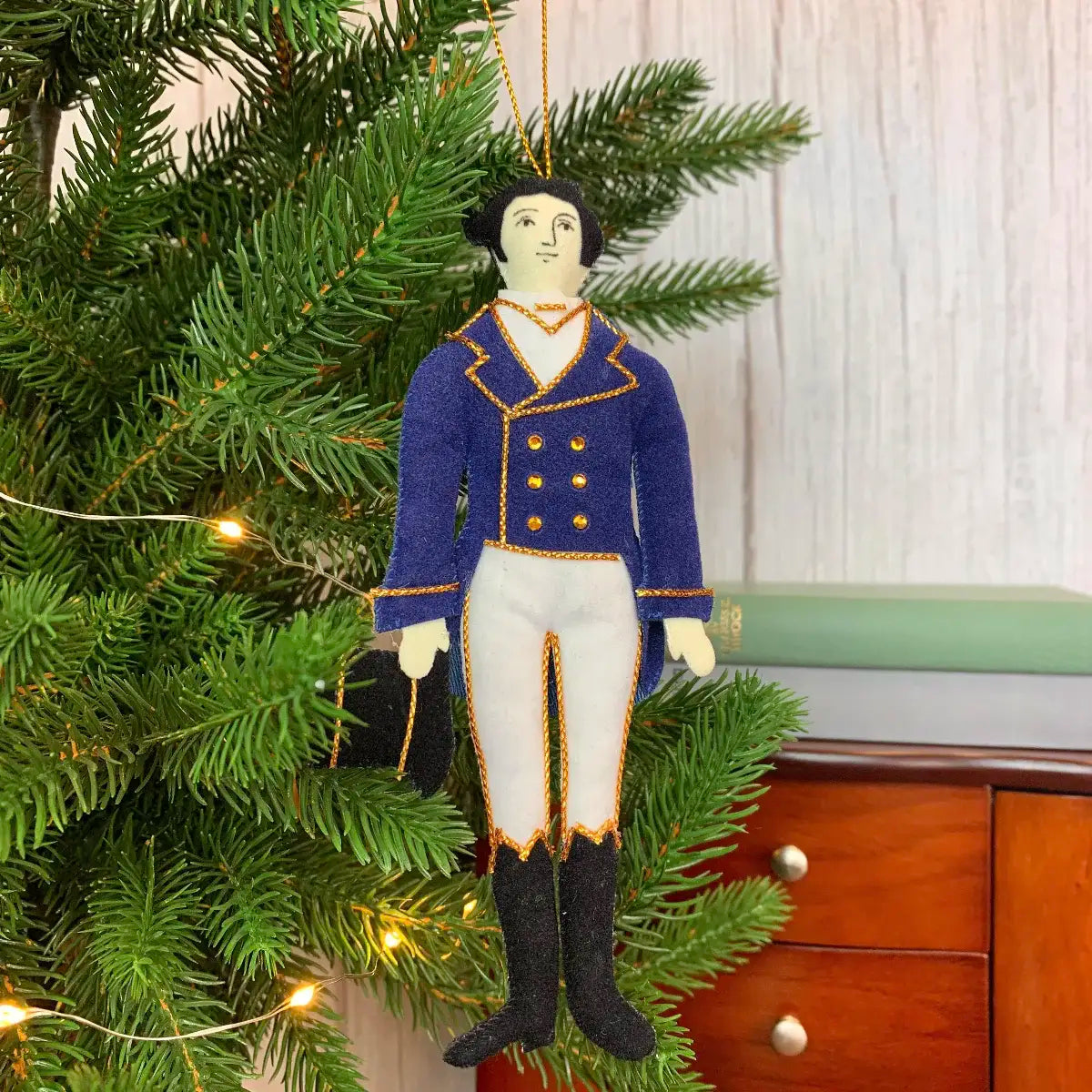 Darcy doll hanging ornament hanging from a christmas tree with lights against a wooden background with a chest of drawers in the background