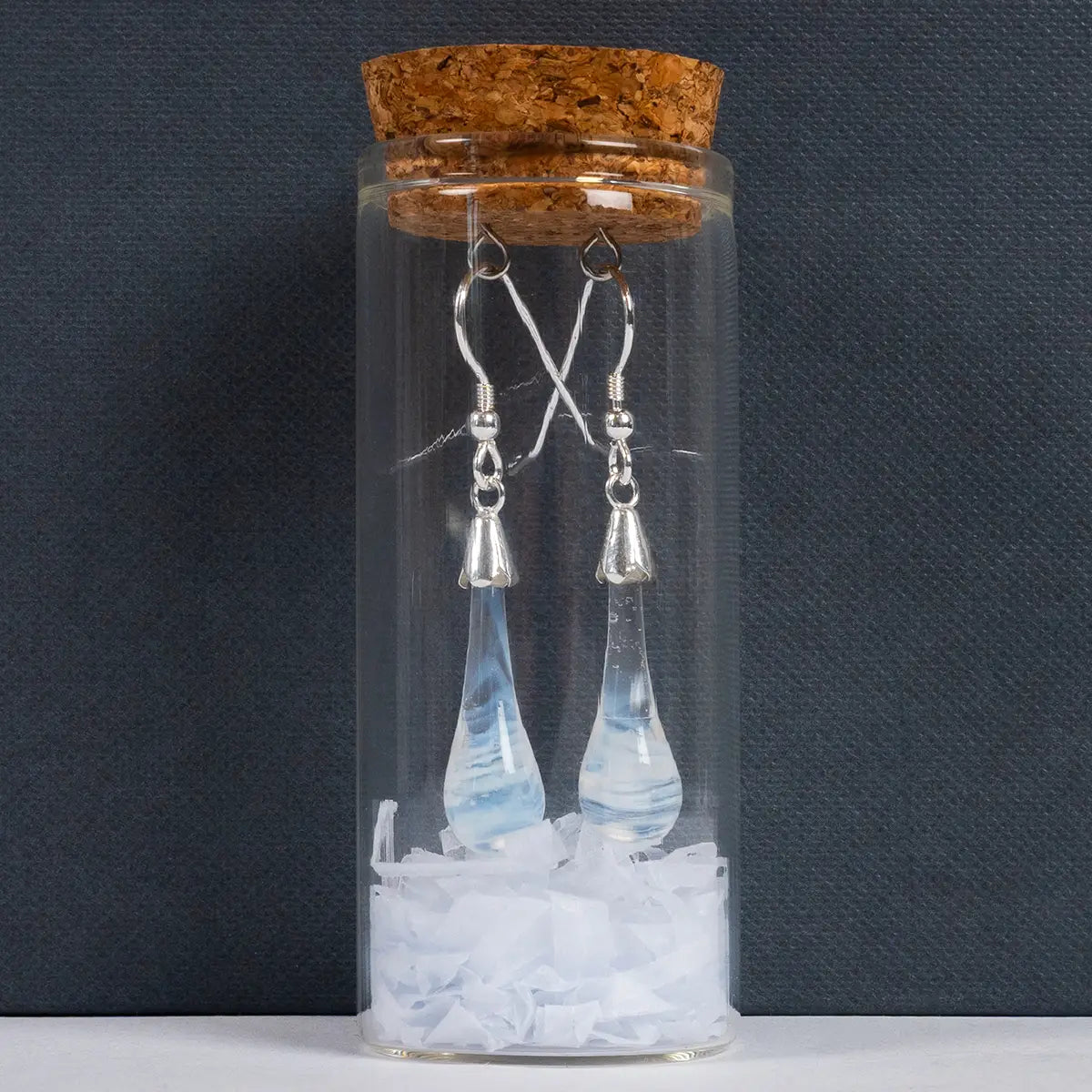 White glass drop earrings with metal details in a glass tube vial with a cork against a dark grey background

