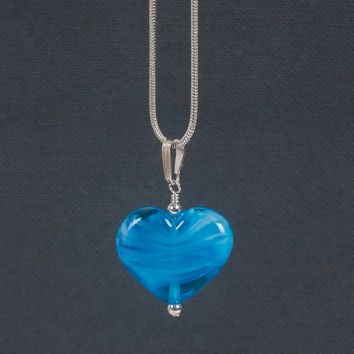 a heart shaped blue glass necklace hanging from a silver chain in front of a grey background