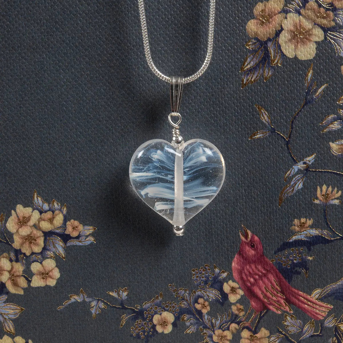 a heart shaped glass necklace on a silver chain against a background printed with birds and flowers