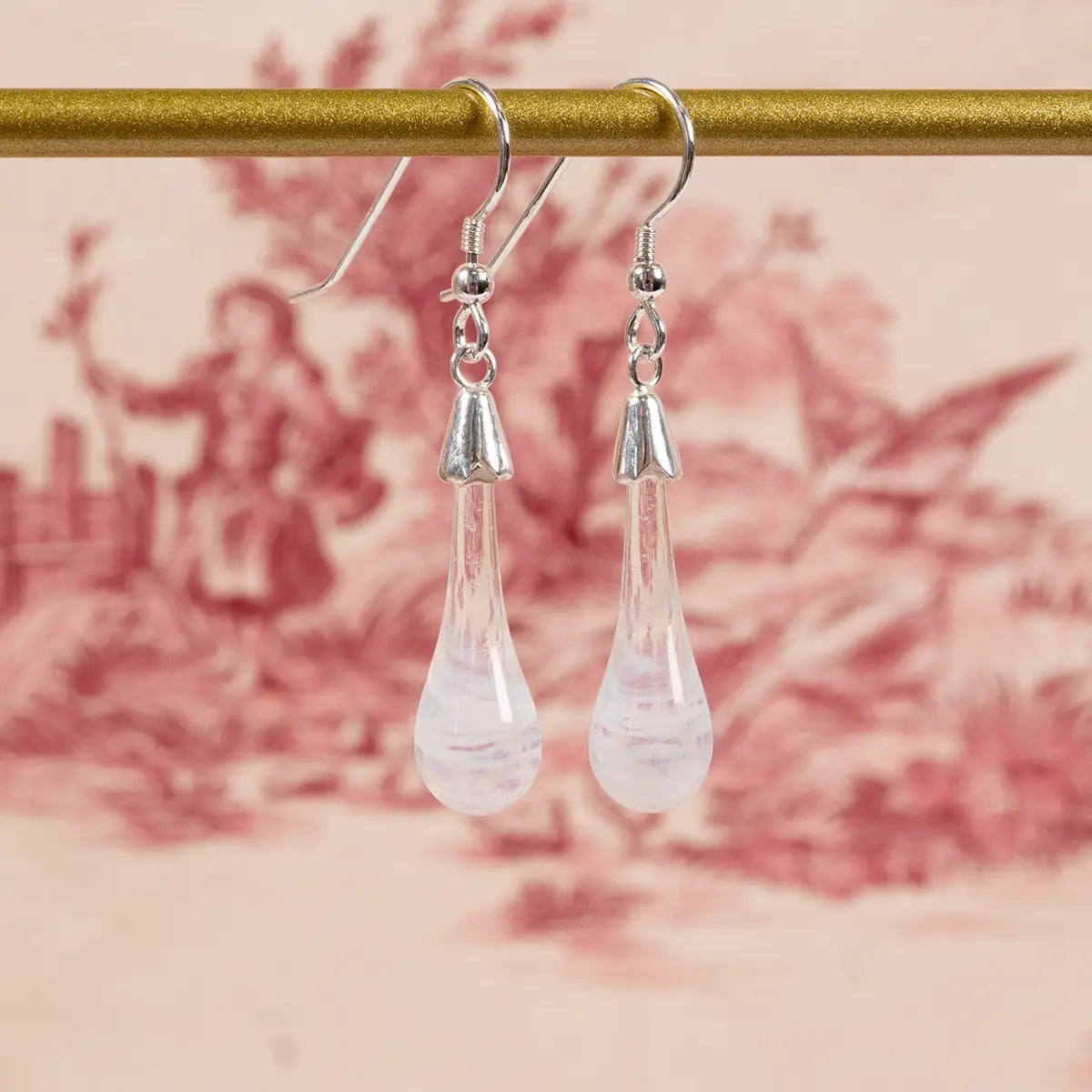 white glass drop earrings with silver details hanging in front of a red patterned sheet