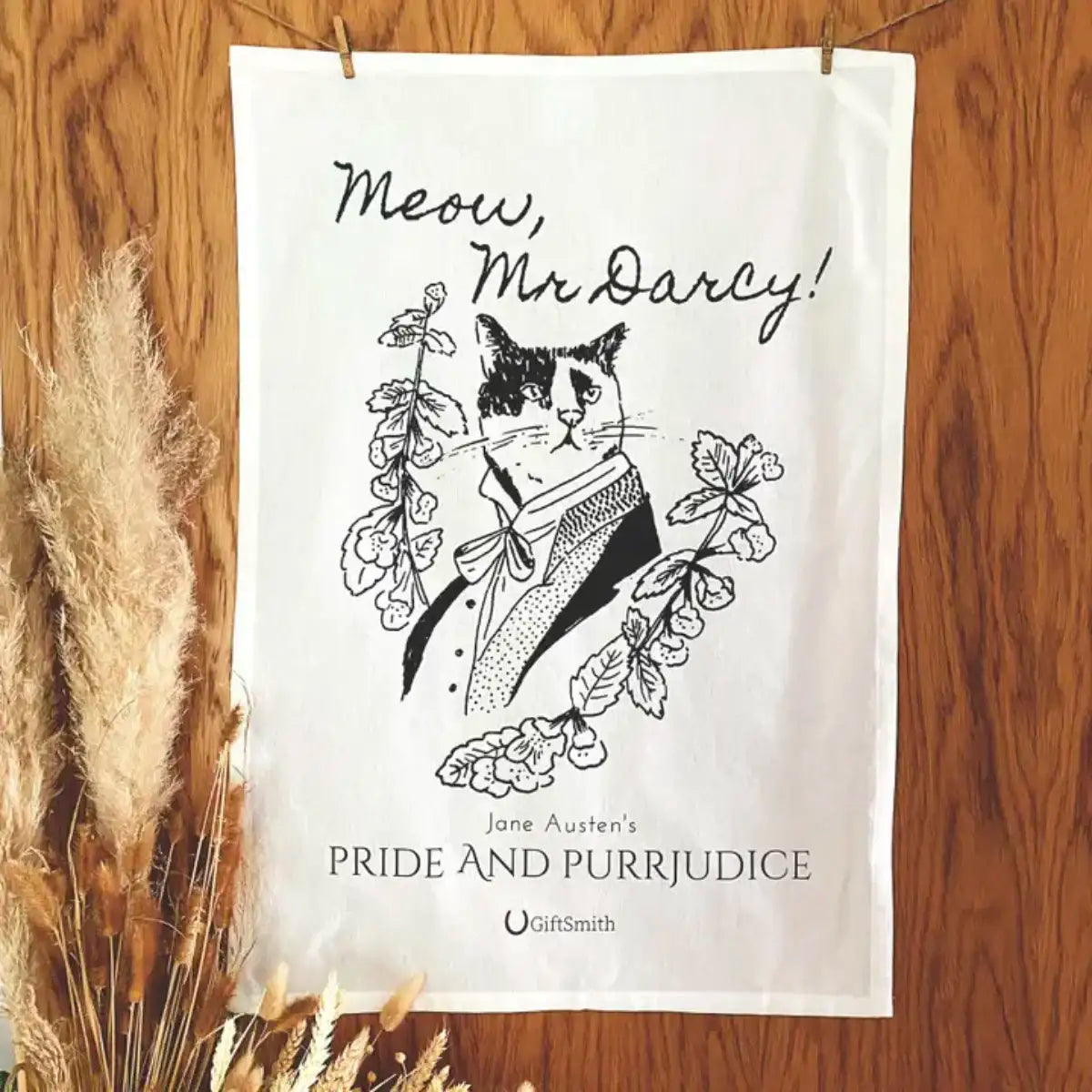 Tea towel showing a line drawing of a cat dressed as Mr Darcy, the towel is hanging from a clothes line in front of a wood effect background with bunch of dried grasses in the foreground.