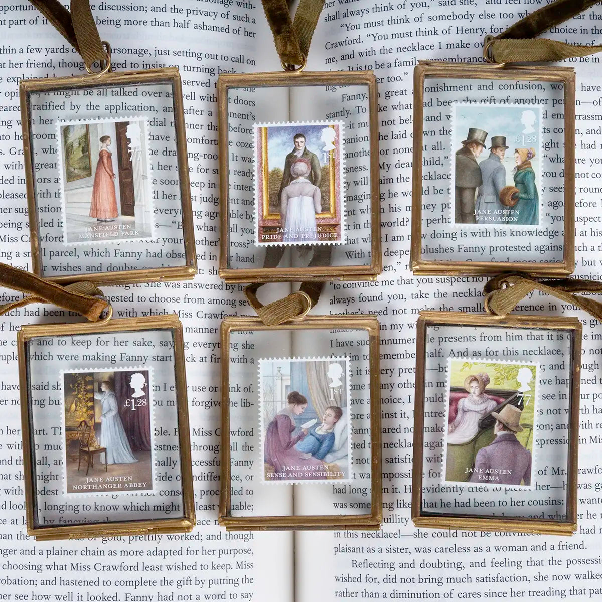 Six Jane Austen themed commemorative stamps in brass cases laid out on a copy of Mansfield Park