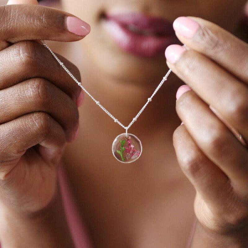 a woman holds the May flower necklace. It is hanging on a silver chain with a a green stem and pink flowers within the pendent. 