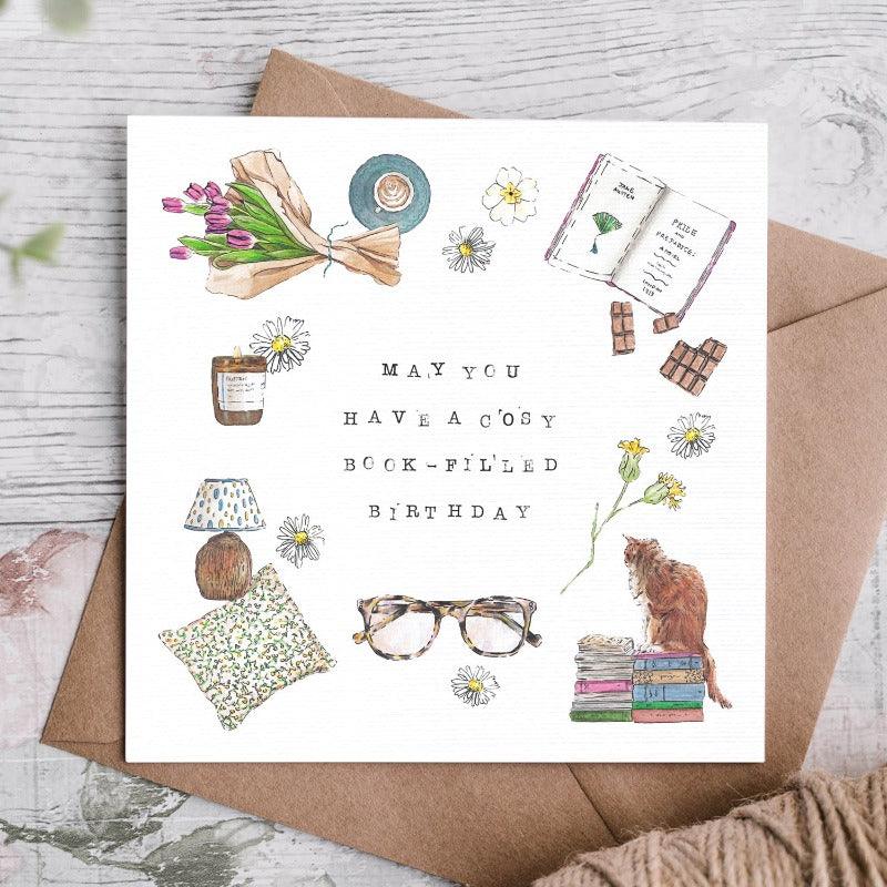 "may you have a cosy book-filled birthday" The card shows, glasses, books, pillows, flowers, chocolate, a candle and a cat illustrated around the words in the centre. 