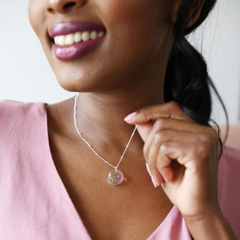 a woman wears the May necklace. It has a green stem and purple flowers set in resin and hanging from a silver chain